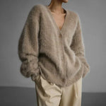 A person wearing a fuzzy, oversized beige cardigan with a V-neck and brown buttons, paired with light beige trousers, set against a plain gray studio background.