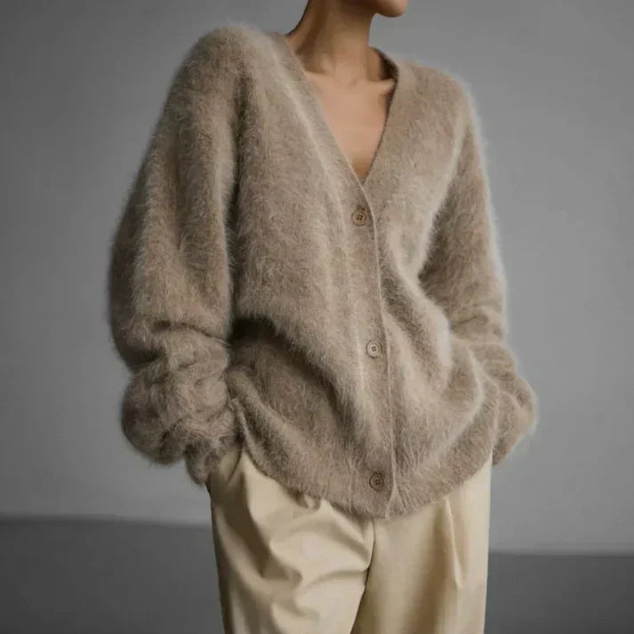 A person wearing a fuzzy, oversized beige cardigan with a V-neck and brown buttons, paired with light beige trousers, set against a plain gray studio background.